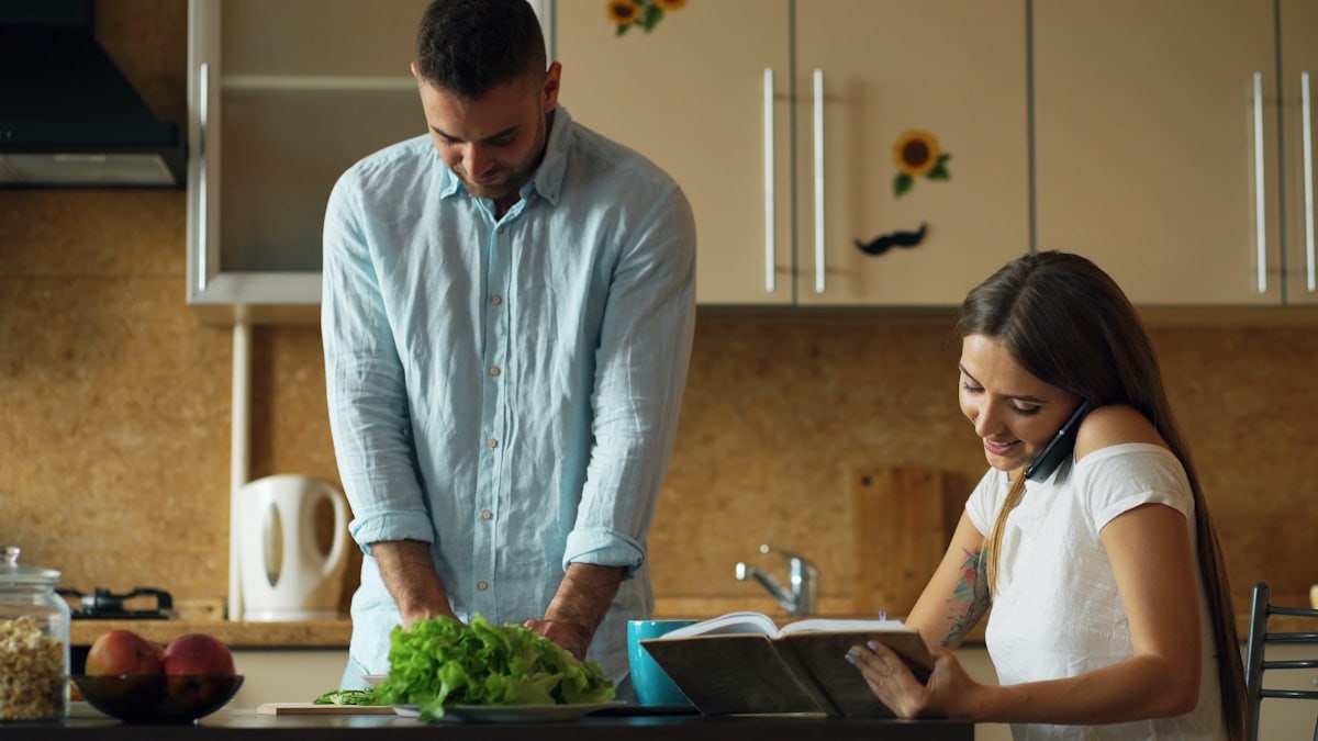 Attractive couple in the kitchen early morning. Beautiful girl chatting phone while her boyfriend cooking breakfast at home