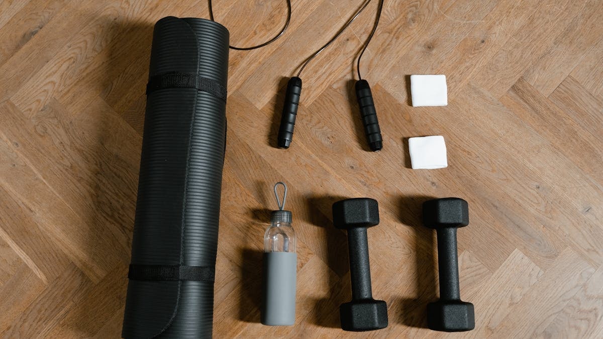 Flat lay of exercise essentials including dumbbells, yoga mat, and water bottle on wooden floor.